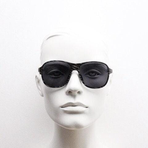 May include: Black aviator sunglasses with a dark, patterned frame. The sunglasses have a classic teardrop shape and dark lenses. The image is a close-up of the sunglasses on a mannequin head against a white background.