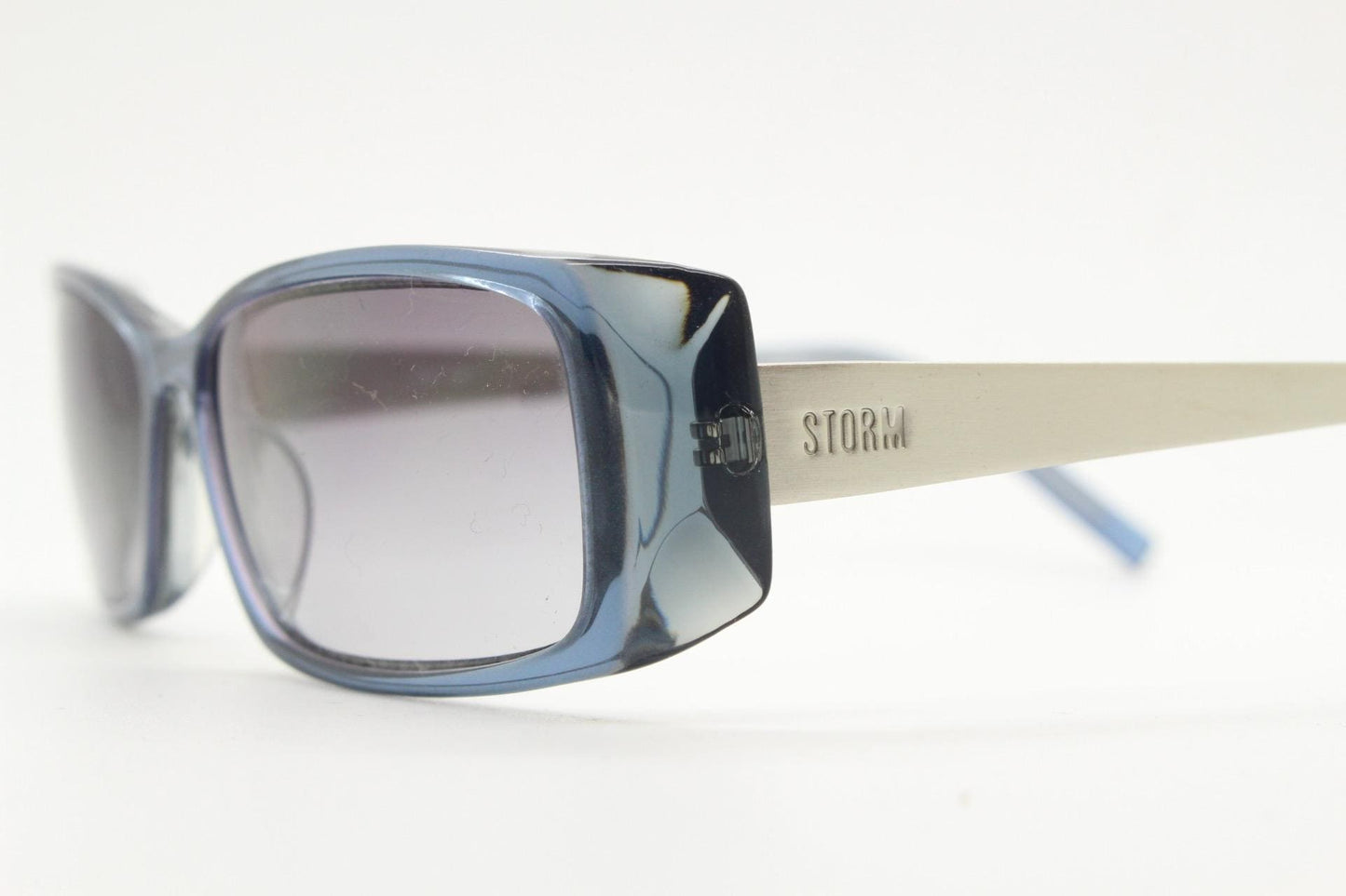 May include: Blue-framed sunglasses with grey lenses. The sunglasses have a rectangular shape with rounded edges. The arms are silver with the word "STORM" printed on the side. The sunglasses are set against a white background.