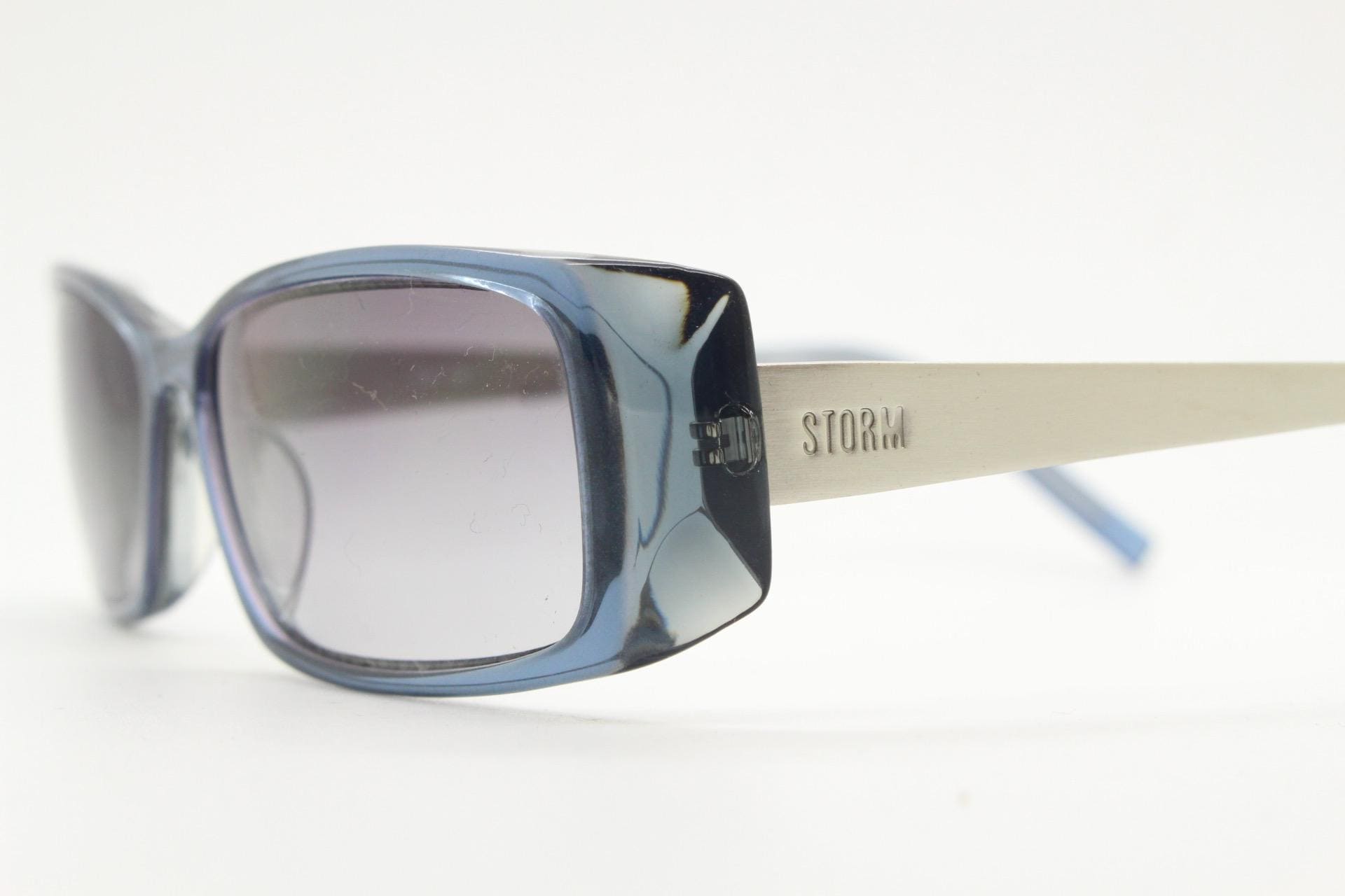 May include: Blue-framed sunglasses with grey lenses. The sunglasses have a rectangular shape with rounded edges. The arms are silver with the word "STORM" printed on the side. The sunglasses are set against a white background.