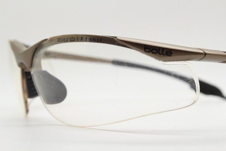 Bolle 90s vintage sports shield sunglasses. Aerodynamic clear lens wrap around half frame design in satin finish with tie string. Cycling