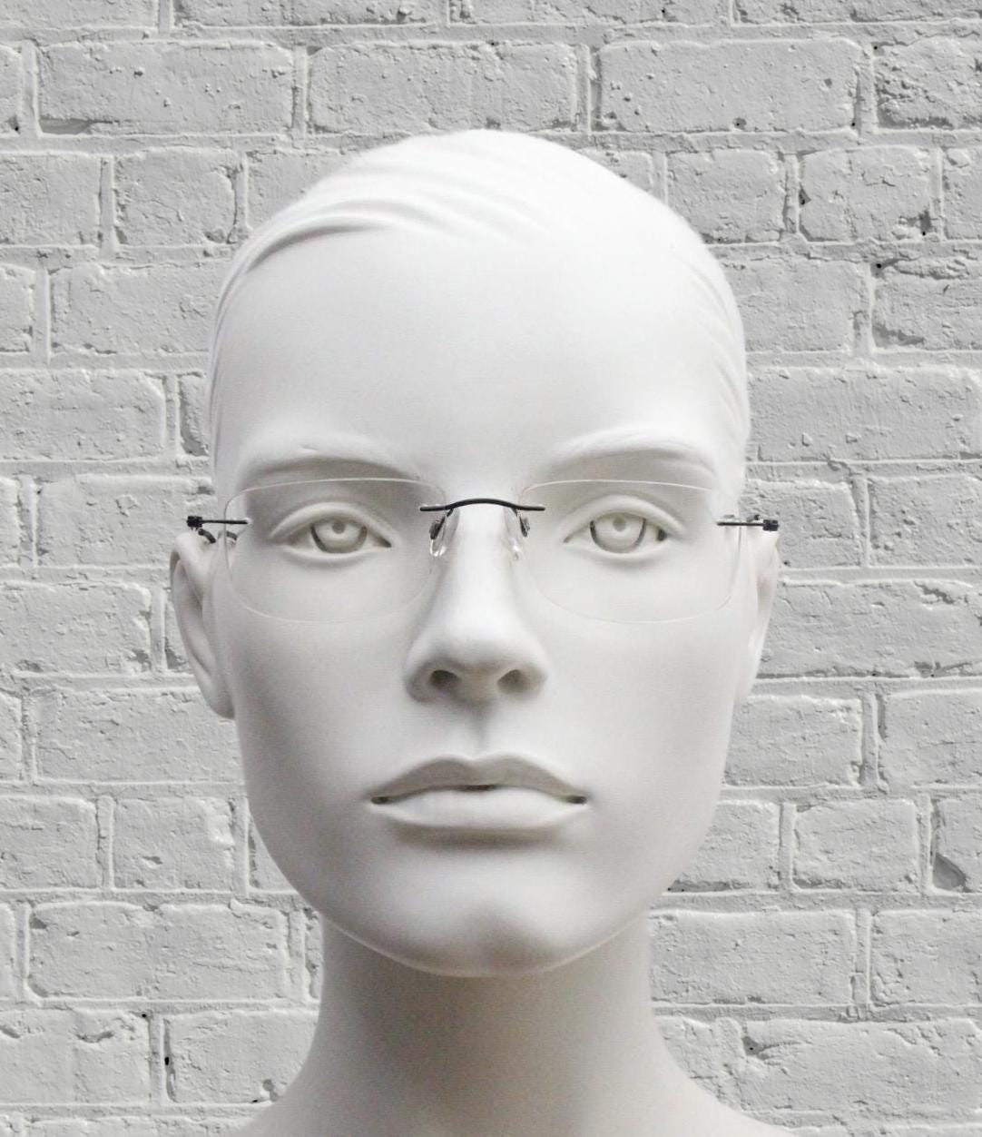 A white mannequin head wearing glasses against a white brick wall background.